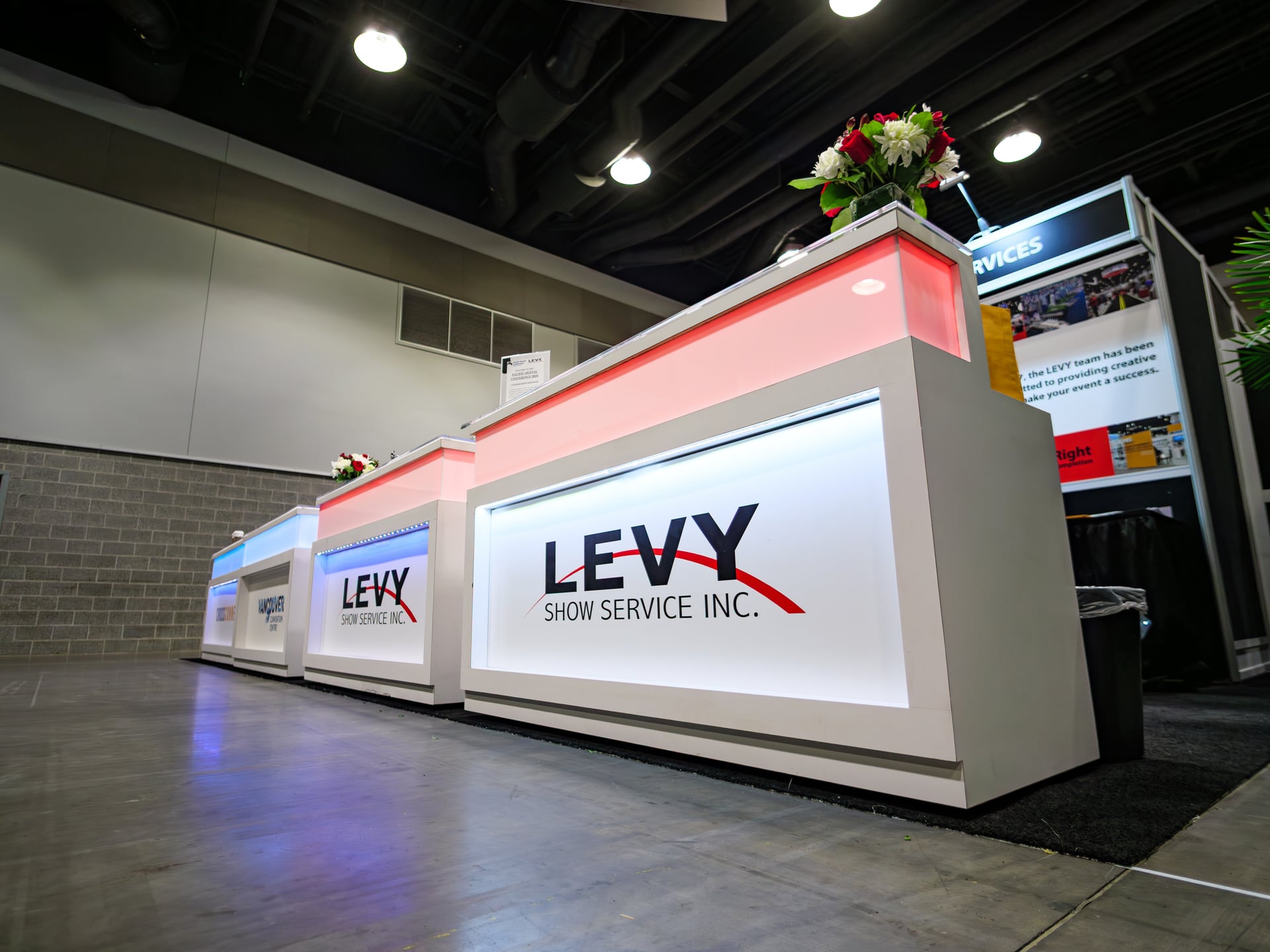 The illuminated LEVY Show Service desk at the Pacific Dental Conference, photographed at night with red accent lighting and a fresh floral arrangement.