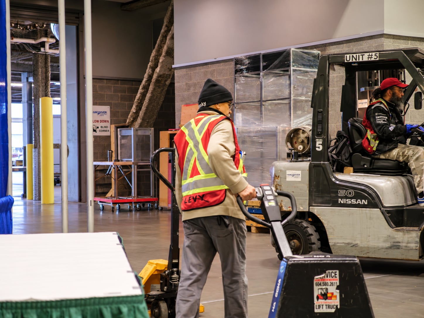 A LEVY crew member in a red safety vest pulls a pallet jack past a forklift on a convention-centre show floor.