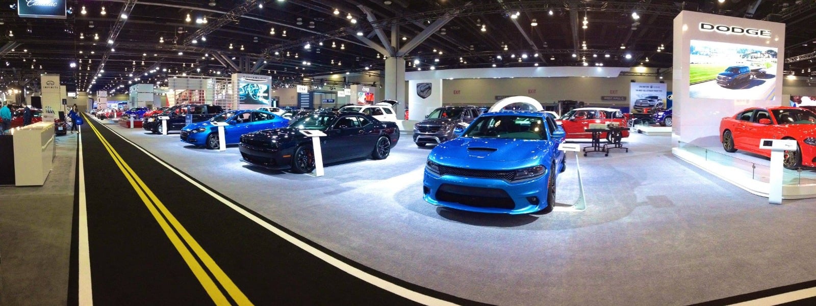 Panoramic view of a finished auto-show floor with multiple manufacturer pavilions including Dodge, Cadillac and Infiniti — vehicles on polished platforms under stadium lighting.