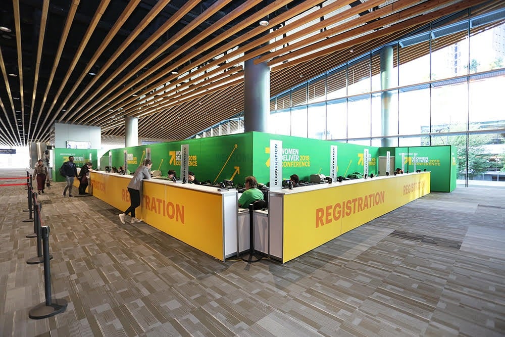 The Women Deliver 2019 Conference registration counter at the Vancouver Convention Centre — a long branded U-shape in green and yellow, with the venue’s signature wood-slat ceiling overhead.